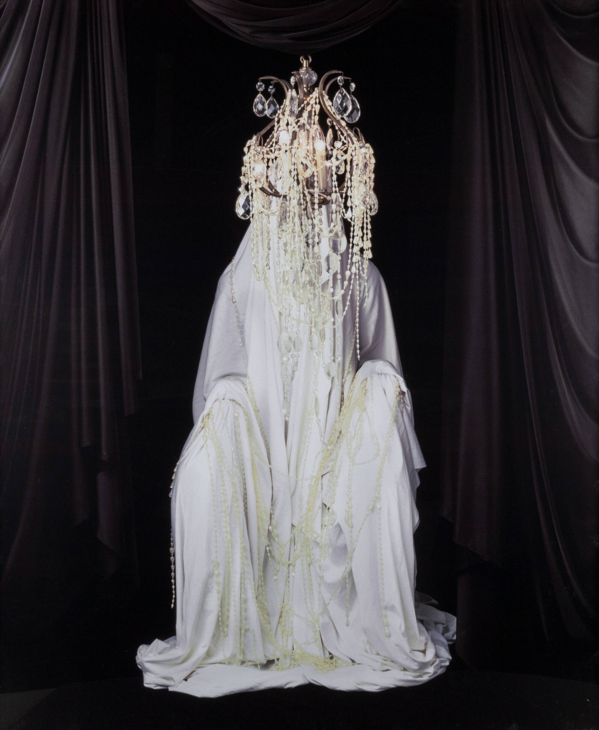 Color photograph of a human figure draped in white fabric against a dark black curtain background with a chandelier on their head.